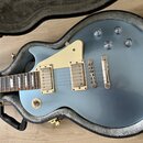 Modrá elektrická kytara v typickém tvaru singlecut, položená v otevřeném kufru s šedým vnitřkem. Nástroj má dva humbuckery, krémový pickguard a čtyři…