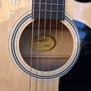 Detail akustické kytary s přirozeným dřevem a černým hmatníkem. Uvnitř otvoru pro zvuk je vidět štítek s nápisy Squier Fender, MODEL NUMBER: SA-105CE…