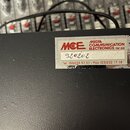 Detail černého zařízení s texturovaným povrchem. Na přístroji je nalepený bílý štítek s červeným logem MCE a nápisem MEDIA COMMUNICATION ELECTRONICS…