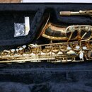 Zlatý lakovaný saxofon v černém kufru. Na čáře nástroje je nápis „ELK SERIES“. Vedle saxofonu je položen munčnípíst s korkem a mouthpiece. Povrch nás…