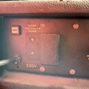 Detail zadní strany černého přístroje v texturovaném obalu. Na panelu jsou nápisy FUSE, 3ASB, 120 VOLTS FUSE a 220V, dále je zde částečně viditelný n…