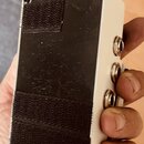 Fotografie zachycuje spodní stranu bílého kytarového pedálu v kovovém šasi. Na spodní části je nalepený černý suchý zips, který vykazuje známky opotř…
