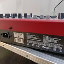 Záběr na zadní červenou část elektronického nástroje s řadou ovládacích knoflíků v horní části. Na šedém štítku je uveden text 230V~ 25W 50/60Hz, FUS…