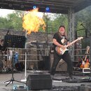 Fotografie zachycuje hudebníky na pódiu. V popředí hraje muž elektrickou kytaru s bílým trupem a hnědým hmatníkem. V pozadí je vidět bicí souprava a…