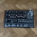 Černý analogový syntetizátor v dřevěném rámu s množstvím potenciometrů, posuvníků a tlačítek. Na spodním panelu jsou vlevo nápis ARTURIA a vpravo MIN…