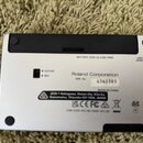 Zadní strana stříbrného elektronického zařízení s černým plastovým panelem. Na spodní straně je nápis Roland Corporation, vedle něj číslo 6342383 a t…