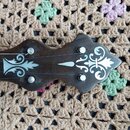 Detail hlavy strunového nástroje z tmavého dřeva s bílými dekorativními ornamenty. Na hlavě jsou čtyři kovové ladičky. Nástroj nemá žádné viditelné z…