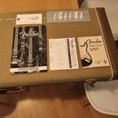 Na hnědém kufr-case s jemným pružkovaným vzorem leží dokumentace a příslušenství. Je zde černý manuál s nápisem Fender, www.fender.com a OWNER'S MANU…