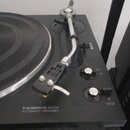 Černý gramofon s kovovým raménkem a černým talířem. Na spodní části šasi je bílý nápis FG-SERVO MOTOR AUTOMATIC TURNTABLE. Vpravo jsou umístěny dva o…