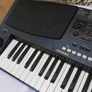 Černý elektronický klavír s bílými a černými klávesami. Na horním panelu je nápis KP300X a tabulky s názvy SONGS a STYLES. Nástroj obsahuje různé ovl…