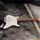 Černá elektrická kytara s bílým pickguardem a hnědým hmatníkem. Na hlavě nástroje je viditelné logo Fender. Kytara má tři snímače, pětistupňový přepí…