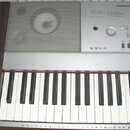 Stříbrošedý digitální klavír s bílými a černými klávesami. V horní části je vlevo mřížka reproduktoru a vpravo ovládací panel s nápisy SONG, STYLE, M…