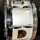 Bílý snare drum s chromovaným kováním. Na koruse je vidět logo Pearl v oválném rámu s nápisem "Pearl" a pod ním menší, hůře čitelný text. Povrch nást…