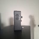 Černý kovový box s dvěma MIDI konektory na přední straně. Vedle konektorů jsou bílé nápisy MIDI IN a OUT/THRU. Na pravé straně zařízení jsou vidět dv…