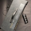 Černý kytarový zesilovač s texturovaným povrchem a plastovou rukojetí. Na zadní straně je viditelný nápis Initial set-up a doprovodný text v angličti…