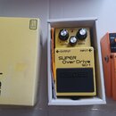 Žlutý efekt pedál v bílém krabici a vedle něj žlutá originální krabice. Na pedálu jsou nápisy BOSS, SUPER Over Drive a SD-1, s ovládacími prvky LEVEL…