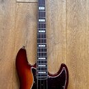 Elektrická basová kytara s trupem v červenohnědém sunburst provedení a hnědým hmatníkem s bílými polohovými značkami. Nástroj má tmavě červený pickgu…