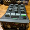 Šedý audio mixér M-CASTER STUDIO s několika ovládacími knoflíky, tlačítky S1 až S4 a malým displejem. Na přední straně je zelený nápis M-CASTER STUDI…