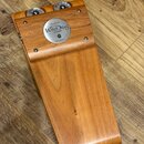 Dřevěný stomp box v přirozené hnědé barvě s viditnou strukturou dřeva. Na horní části jsou umístěny dvě kovové pružiny a kulaté stříbrné logo s nápis…