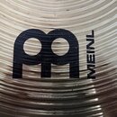 Detailní záběr na povrch kovového činely s koncentrickými drážkami. Uprostřed je černé logo ve tvaru stylizovaného písmene M a vedle něj svisle napsa…