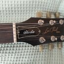 Detail hlavy černé elektrické kytary s hnedým hmatníkem. Na hlavě jsou bílé nápisy „Studio“, „Les Paul MODEL“ a logo „Gibson“. Povrch je glossy, s vi…