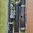 Tři části dřevěného dechového nástroje v tmavě hnědé barvě, umístěné v otevřeném kufru s zeleným polstrováním. Nástroj má kovové klapy a prstence v h…
