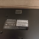 Fotografie zachycuje spodní část černého zařízení s přilepeným bílým štítkem. Na štítku je uvedeno logo a název KOMPLETE, model Komplete Kontrol S61…