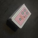 Bílá krabička s růžovým panelem, na kterém je nápis FUZZ a logo Fender. V pravé části panelu je obrázek postavičky Hello Kitty a tři stylizované ovlá…