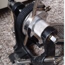 Detailní záběr na černý kovový mechanismus bicího pedálu s kovovými šrouby a spojovacími prvky. Na mechanismu je vidět černý texturovaný řemen a část…