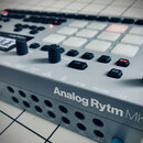 Stříbrošedý sampler/drum machine s bílými podsvícenými pady a černými ovratovými knoflíky. Na přední straně je bílý nápis Analog Rytm MKII. Zařízení…