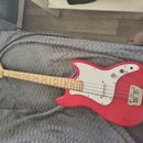 Červená elektrická basová kytara s bílým pickguardem a hnedým hmatníkem. Na hlavě je viditelné logo Squier. Nástroj vykazuje známky běžného používání…