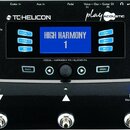Černý procesor efektů s modrým displejem, na kterém je nápis HIGH HARMONY 1. Na horní části jsou loga TC-HELICON a play ACOUSTIC. Zařízení obsahuje n…