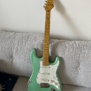 Světle zelená elektrická kytara s bílým pickguardem a javorovým hmatníkem. Na hlavě je viditelné logo Fender. Lak na těle vykazuje výrazné opotřebení…