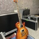 Elektrická kytara s trupem v červeně-žlutém sunburst provedení a viditnou strukturou dřeva. Na hlavě hmatníku je bílé logo PRS. Nástroj má dva humbuc…