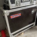 Dvě černé transportní bedny s hliníkovými hranami a kovovými zapojovacími prvky. Horní bedna má nápisy „R-STICK PROFESSIONAL DRUMSTICKS“, „www.rstick…
