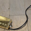 Zlatý humbucker s kovovým krytem a šesti viditelnými šrouby v polosičce. K snímači vede černý kabel s odhalenými koncovými vodiči a bílým papírovým š…