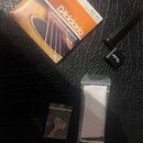Na černém texturovaném povrchu leží balení kytarových strun D'Addario PHOSPHOR BRONZE EJ15 Extra Light Gauge 010-.047. Vedle nich je černý kytarový k…