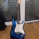 Modrá elektrická kytara s bílým pickguardem a javorovým hmatníkem. Na hlavě je viditelné logo Fender. Kytara stojí na černém stojanu a vedle ní je če…
