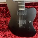 Černá elektrická kytara s matným povrchem v červeném pouzdře. Nástroj má dva humbuckery, na kterých je čitelný nápis emg. Na ovládacím knoflíku je ná…