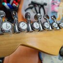 Detail hlavy světlé dřevěné kytary s šesti chromovanými ladicími mechanismy. Na vřetenech ladíček jsou bílé kruhové značky s černým symbolem připomín…