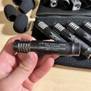 Fotografie zachycuje několik černých kondenzátorových mikrofonů s mřížkovým vrchem. Na těle drženého mikrofonu je bílý nápis: 9-52 VOLT REMOTE POWER…