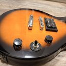 Detail těla elektrické kytary s barevným přechodem od oranžové k černé. Nástroj má dva humbuckery, kovový most a tři ovládací prvky: dva otočné knofl…