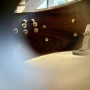 Detailní pohled na tmavě hnědý lakovaný dřevěný povrch nástroje. V horní části jsou vidět různé šrouby a vruty zapuštěné do dřeva, některé z nich jso…