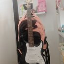 Černá elektrická kytara s bílým pickguardem a světle hnědým hmatníkem. Na hlavě nástroje je viditelné logo Squier. Kytara stojí na stojanu před růžov…
