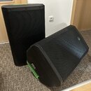 Dva černé reproduktory s kovovou mřížkou. Na pravém reproduktoru je viditelné zelené kulaté logo s písmeny „JB“. Jeden reproduktor stojí vzpřímeně, d…