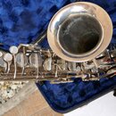 Stříbrný saxofon v modrém polstrovaném kufru. Je vidět detail čarovnice a část klapového mechanismu s perleťovými stisky. Povrch nástroje vykazuje dr…