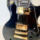 Černá elektrická kytara s gold hardwarem a bílým bindingem. Na těle jsou dva zlaté humbuckery, zlatý kobylník a čtyři černé oválné knofíky. V horní č…