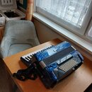 Modrá akordeon s mramorovaným povrchem a bílými klávesami. Na horní části je viditelné logo "Hohner". Nástroj je vybaven černými popruhymi a bílými b…