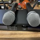 Dva černé bezdrátové mikrofony s kovovými mřížkami leží na černém přijímači. Na přední straně přijímače jsou tlačítka s nápisy SET, AUTO a ADL oraz n…