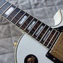 Bílá elektrická kytara s tmavým hmatníkem a perleťovými vložkami. Na těle je umístěn přepínač s nápisy TREBLE a RHYTHM. Nástroj má zlaté potenciometr…